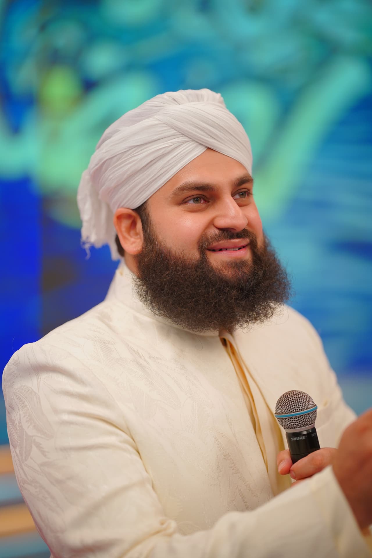 Hafiz Ahmed Raza Qadri wearing a white turban and patterned sherwani, holding a microphone
